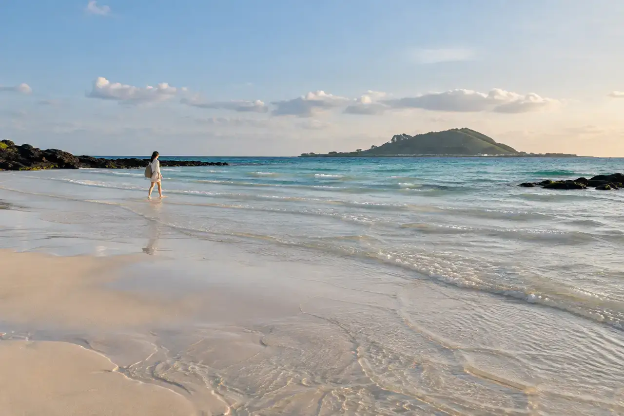 Pale sand, shallow blue water and a low island offshore on the west Jeju coast.