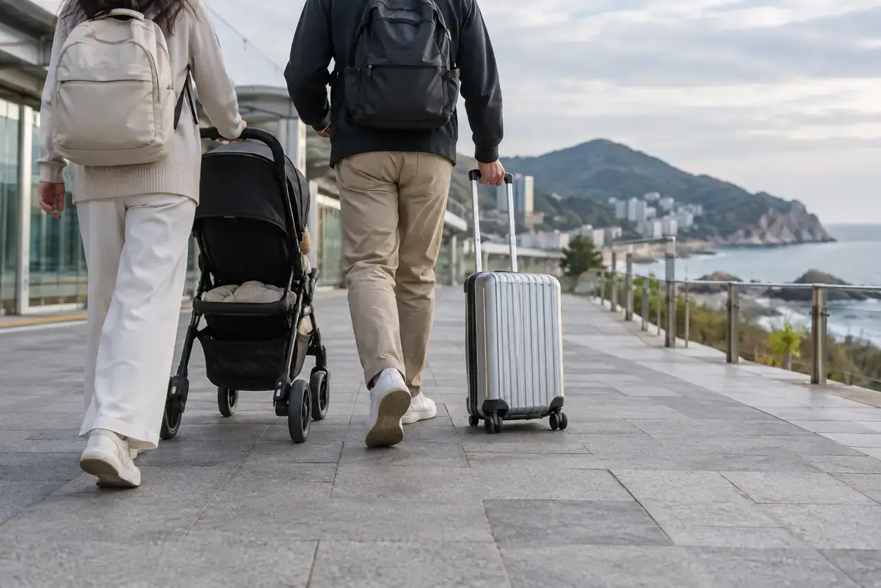 Two travelers with a stroller and rolling suitcase on a coastal pedestrian path in Busan.