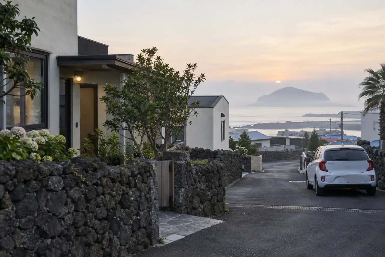 A quiet dawn street in Jeju City with a volcanic-stone wall and an unbranded rental car, illustrating a first-night base after a late arrival